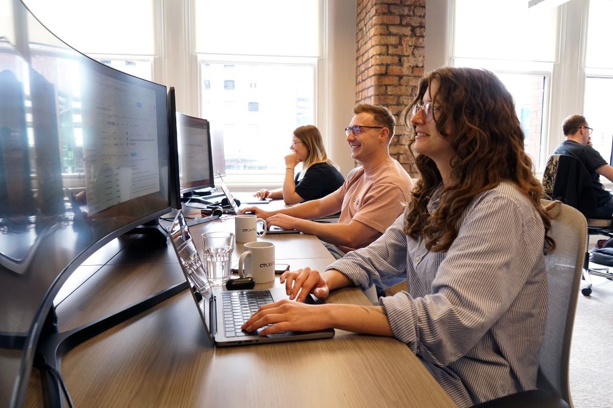 three people sat at desks typing on their laptops and smiling